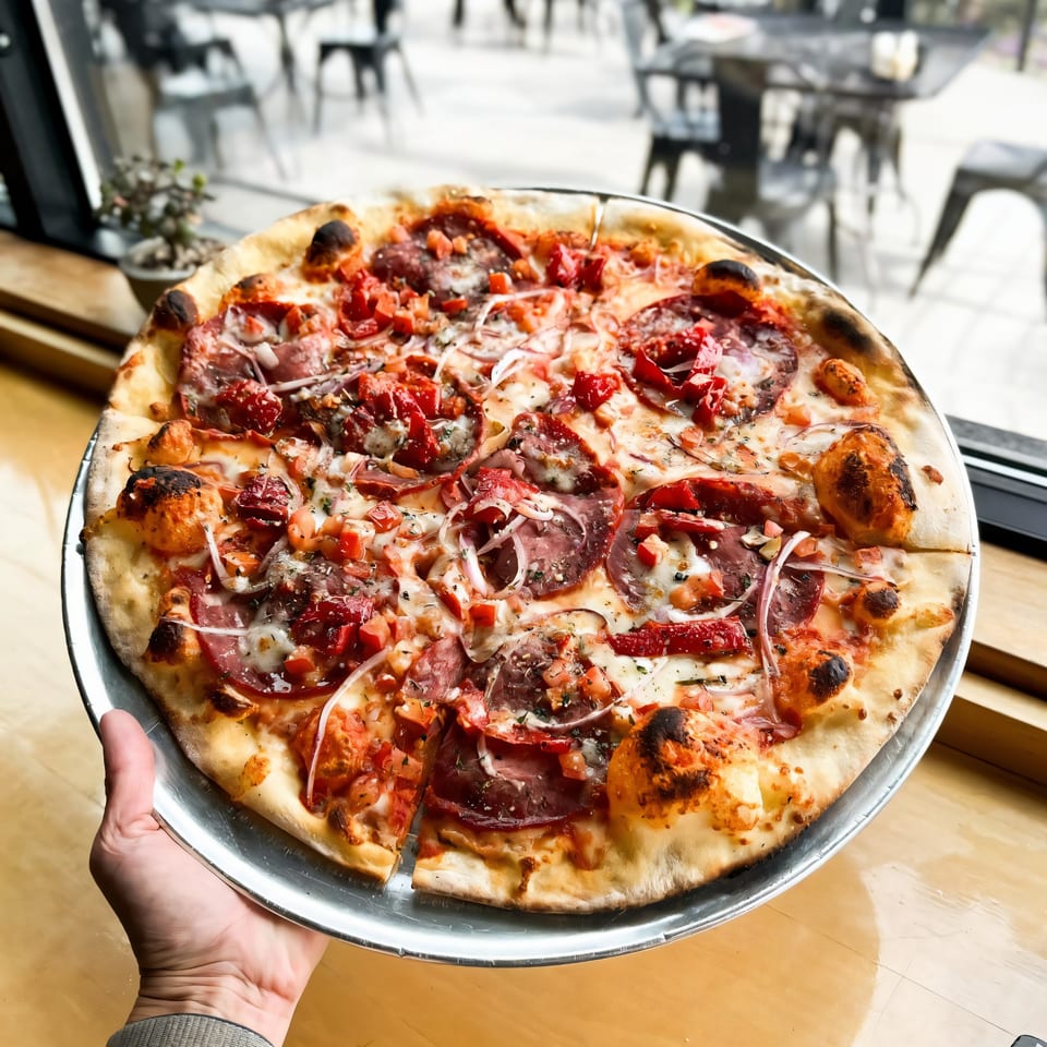 Hand holding a Versalia Pizza with pepperoni, red onions, and diced tomatoes, by a window.