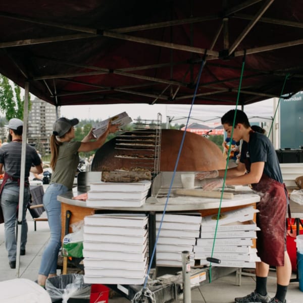 Versalia Pizza staff preparing pizzas at an outdoor event with a large wood-fired oven.