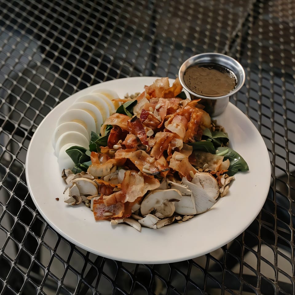 A Versalia Pizza salad with sliced hard-boiled eggs, mushrooms, crispy bacon, and dressing on a white plate.