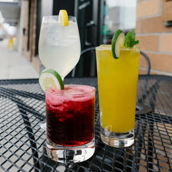 Three colorful Versalia Pizza cocktails, red, yellow, and clear, garnished with citrus and mint on a patio table.