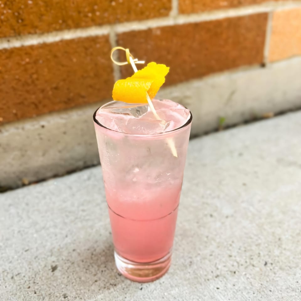 A pink cocktail with ice and an orange peel garnish sits on a concrete surface.