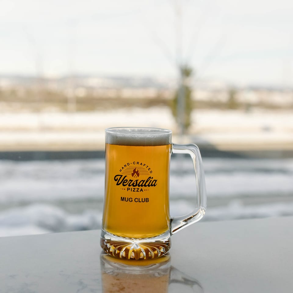 A Versalia Pizza Mug Club beer stein filled with golden beer and foam, against a snowy window.