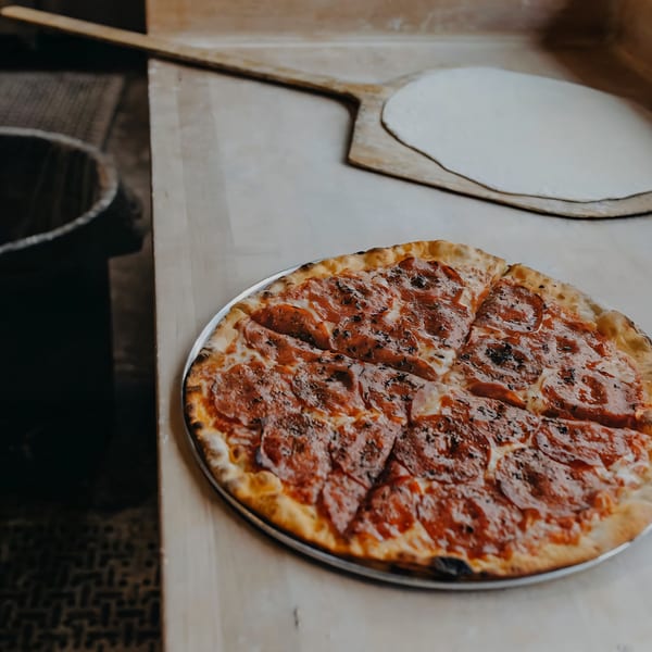 A pepperoni pizza on a metal tray next to a pizza peel with raw dough.