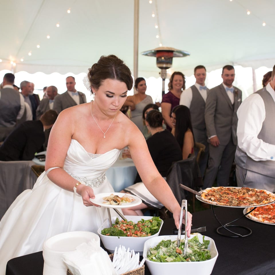A bride in a white gown serves herself pizza and salad from a buffet at a wedding reception.